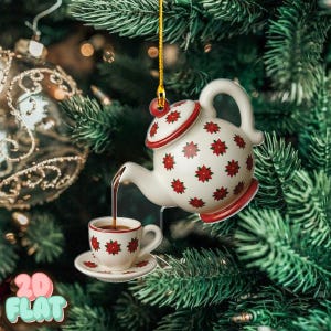 May include: A white ceramic teapot and teacup Christmas ornament with red floral detailing. The teapot is pouring a dark liquid into the teacup. The ornament is suspended in front of a Christmas tree.