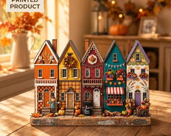 May include: A 2D flat printed product featuring a row of five colorful miniature houses on a wooden base. The houses are decorated with autumn themes including pumpkins, fall foliage, a scarecrow, a wreath, and a black cauldron labeled Spooky Treats.