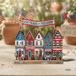 May include: A patriotic village scene with five miniature houses decorated with American flags and festive details. A banner displays "250 YEARS OF FREEDOM". The houses are painted in red, white, and blue, celebrating Independence Day.