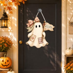 May include: A hanging door sign shaped like a white ghost with black eyes, featuring a large patterned bow on its head, two small floral and plaid patches on its body, and a small flag held in one hand with the word WELCOME printed on it.