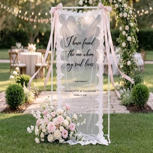 Peut inclure: Panneau de mariage blanc avec un tissu en dentelle transparent. Le panneau affiche le texte "J'ai trouvé celui que mon âme aime." et les noms Charlotte & Theodore, avec la date du 12 octobre 2026. Des rubans roses et des fleurs ornent le panneau.