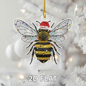 May include: A clear acrylic Christmas ornament shaped like a bee, decorated with a Santa hat and string lights. The bee has black and yellow stripes and the name "Sophia" is written on the hat. The ornament is hanging from a gold string.