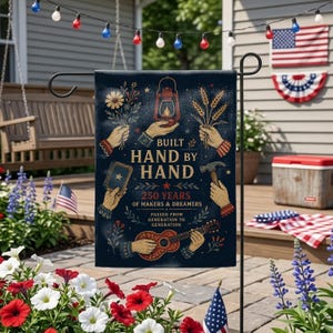 May include: A navy blue garden flag with the text "BUILT HAND BY HAND" and "250 YEARS OF MAKERS & DREAMERS." The flag features illustrations of hands holding a lantern, flowers, wheat, a hammer, a book, and a guitar. The flag is displayed outdoors.