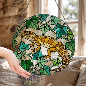 May include: Circular stained glass window art depicting a gecko in yellow, brown, and orange, framed by green ivy leaves. The artwork hangs from a chain, designed as a decorative piece for a window or wall.