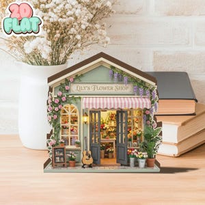 Puede incluir: Una maqueta en miniatura de una floristería con el texto "Lily's Flower Shop". La tienda tiene una fachada verde, un toldo a rayas y está decorada con flores y enredaderas. Una guitarra se apoya en la tienda y también se ve una pequeña pizarra.