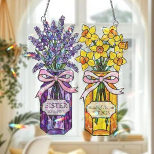 May include: Two decorative glass jar ornaments hanging from chains. One jar features lavender and the text "SISTER EST.1997". The other jar has yellow daffodils and the text "Maid of Honor EST.2026". Both jars have pink bows.