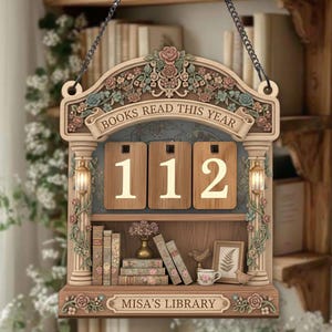 May include: Wooden wall hanging with the text "BOOKS READ THIS YEAR" and the number "112". The decorative piece features a shelf with miniature books, a vase, a teacup, and the text "MISA'S LIBRARY". It is adorned with floral and scrollwork details.
