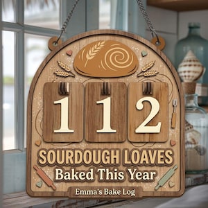 May include: Wooden sign featuring a bread loaf illustration, wheat stalks, and the numbers "112." Text reads "Sourdough Loaves Baked This Year" and "Emma's Bake Log." Includes rolling pin and heart accents. Hanging chain at the top.