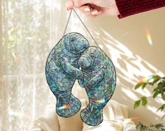 May include: A decorative hanging featuring two manatees embracing, crafted from mosaic-style glass. The manatees are primarily blue and green, with a chain for hanging. The image is held by a person's hand.