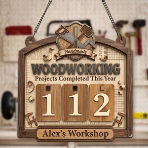 May include: Wooden woodworking sign with the text "Woodworking Projects Completed This Year" and the number "112". The sign also includes the text "Handmade" and "Alex's Workshop". The sign is hanging from a chain.