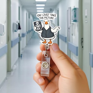 Pode incluir: Um porta-crachá de desenho animado de um ganso segurando um raio-x, com o texto "IDK I JUST TAKE THE PICTURE". O ganso está vestindo um colete. O porta-crachá tem uma alça de plástico transparente e um botão de metal.