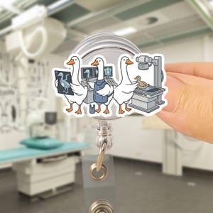 Könnte beinhalten: Ein Ausweis-Rollhalter mit einem Cartoon-Design von drei weißen Gänsen in einer medizinischen Umgebung. Eine Gans hält ein Röntgenbild, eine andere trägt eine blaue Schürze und eine dritte untersucht eine Ente. Der Hintergrund ist ein verschwommenes medizinisches Büro.