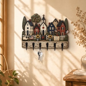 May include: A wall-mounted key holder featuring a decorative scene of a small village with houses, a church, trees, and a white picket fence. The base has a distressed wood finish with six black metal hooks, one holding a set of silver keys.