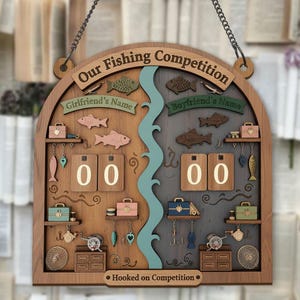 Puede incluir: Letrero de competición de pesca de madera con el texto "Our Fishing Competition". El letrero presenta peces decorativos, cajas de aparejos y un diseño de río. Incluye espacios para rastrear puntuaciones y está diseñado para colgarse.