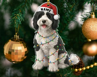 Puede incluir: Adorno navideño con un perro blanco y negro con un gorro de Papá Noel con la palabra "NAME" y luces de colores. El adorno cuelga de un árbol de Navidad con adornos dorados y verdes.
