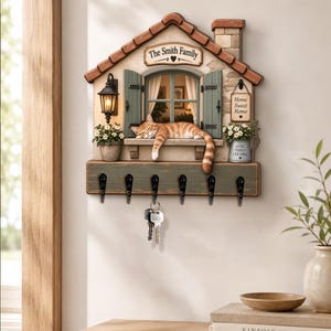 Puede incluir: Un llavero de pared con forma de casa con techo marrón, un gato atigrado naranja durmiendo en el alféizar de una ventana y contraventanas verdes. Incluye una linterna, dos plantas decorativas en macetas y el texto The Smith Family y Home Sweet Home.