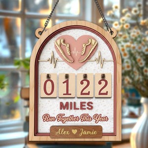 Könnte beinhalten: Holzschild mit hellbraunem Rahmen und Kette zum Aufhängen. Das Schild zeigt ein rosa Herz mit Laufschuhen, eine Herzschlaglinie und den Text "0122 Miles Run Together This Year." Enthält die Namen Alex und Jamie.