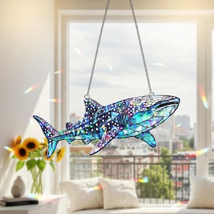 May include: A stained glass whale shark suncatcher, suspended by a silver chain. The shark features a mosaic design with blue, green, and purple hues, and white spots. The background shows a cityscape and a window with sunlight.