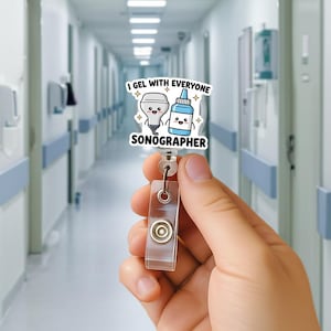 Könnte beinhalten: Ein Ausweis-Rollhalter mit einem Cartoon-Design, das ein Sonogrammgerät und eine Flasche Gel zeigt, mit dem Text "I GEL WITH EVERYONE" und "SONOGRAPHER". Die Rolle ist transparent und hat ein einziehbares Kabel.