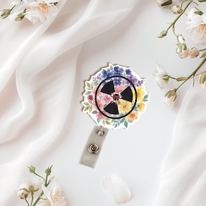 Könnte beinhalten: Ein floraler Ausweis-Aufroller mit einem schwarzen Strahlungssymbol in der Mitte, umgeben von einem Aquarellkranz aus bunten Blumen. Der Ausweis-Aufroller hat ein graues, einziehbares Kabel und einen Metallclip.