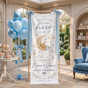 May include: A vertical white fabric banner for a baby shower featuring a sleeping teddy bear on a crescent moon, clouds, and stars. The text reads: Our little star is almost here, Welcome to Emma & James' Baby Shower, August 16, 2026.