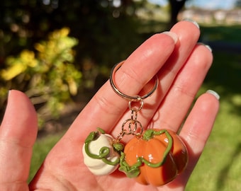 May include: A gold-colored keychain featuring two miniature pumpkins. One pumpkin is orange, and the other is white, both with green vines. The keychain is held in a hand against a blurred green background.