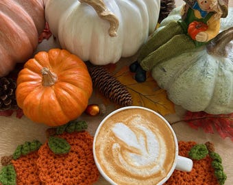May include: A fall-themed still life featuring a white coffee cup with latte art, surrounded by decorative pumpkins in orange, white, and green. Crochet pumpkin coasters and fall foliage complete the autumn arrangement.