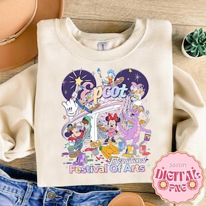 May include: Cream-colored sweatshirt featuring a colorful graphic of Disney characters painting at Epcot's International Festival of Arts. The design includes Mickey Mouse, Minnie Mouse, Donald Duck, and a purple dragon. The text "Epcot" and "International Festival Of Arts" are also visible.