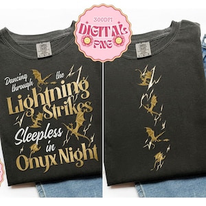 Op de afbeelding: Twee antracietgrijze t-shirts met gouden afbeeldingen. Het linker t-shirt toont de tekst Dancing through Lightning Strikes Sleepless in Onyx Night met illustraties van draken en bliksemschichten. Het rechter t-shirt toont een rij draken.