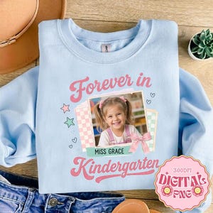 Op de afbeelding: Een lichtblauw sweatshirt met ronde hals en een centrale opdruk met de tekst "Forever in Kindergarten" en "Miss Grace". Het ontwerp bevat een foto van een jong kind, roze ruitjeskaders, kleine sterretjes, hartjes en een roze strik.