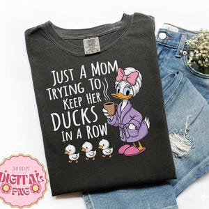 Op de afbeelding: Donkergrijs t-shirt met de tekst "JUST A MOM TRYING TO KEEP HER DUCKS IN A ROW." Toont een cartoon eend in een badjas met een mok, met drie eendjes. Het t-shirt wordt getoond met een spijkerbroek.