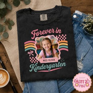 Może przedstawiać: Czarna koszulka z grafiką zawierającą napis "Forever in Kindergarten" oraz "MISS OLIVIA". Projekt obejmuje centralną ramkę ze zdjęciem uśmiechniętego dziecka, kolorowe łuki tęczy, wzory w szachownicę oraz ikonę małej kokardki.