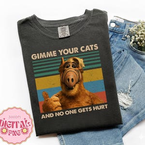 Puede incluir: Camiseta gris oscuro con un diseño gráfico retro del personaje Alf, con el texto "GIMME YOUR CATS AND NO ONE GETS HURT." El diseño incluye un gesto de pulgar hacia arriba y rayas coloridas.