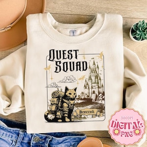 Op de afbeelding: Een crèmekleurige sweater met ronde hals en een grafische print met de tekst "QUEST SQUAD" en "Broccoli". De illustratie toont een kat met een rugzak voor een groot kasteel, omringd door wolken en vliegende draken.