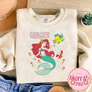May include: Cream-colored sweatshirt featuring a colorful graphic of Ariel from Disney's The Little Mermaid. The design includes Ariel with red hair, a teal tail, and Flounder. The text "The Little Mermaid" is above the image. The sweatshirt is folded on a wooden surface.