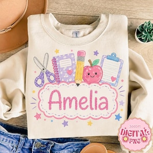 May include: A cream-coloured crewneck sweatshirt featuring a graphic print of school supplies including purple scissors, a lavender notebook, a yellow pencil, a smiling red apple, and a blue clipboard. The name Amelia is printed in pink.
