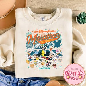 Könnte beinhalten: Cremefarbenes Sweatshirt mit einem farbenfrohen Grafikdesign von Disney-Figuren beim Laufen. Der Aufdruck enthält den Text "Walt Disney World Marathon Weekend 2026" und "runDisney". Mickey Mouse, Minnie Mouse, Donald Duck und Daisy Duck sind ebenfalls abgebildet.