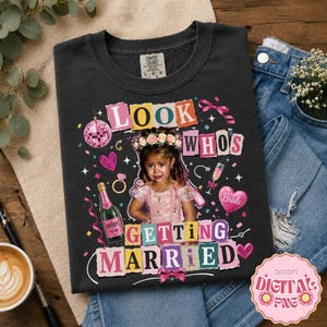 May include: A charcoal gray t-shirt featuring a graphic print of a child wearing a floral crown. The text reads "LOOK WHO'S GETTING MARRIED" in colorful, collage-style letters. The design includes a disco ball, champagne bottle, and hearts.