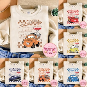 Può includere: Una collezione di felpe beige con disegni di auto dei cartoni animati. Una felpa ha "Tow Matter" e un carro attrezzi dei cartoni animati. Altre felpe presentano grafiche di auto "McQueen", "Luigi", "Shot Rod", "Hudson" e "Sally Carrera".