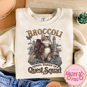 Op de afbeelding: Een crèmekleurig sweatshirt met ronde hals en een afbeelding van een bruine cyperse kat met een rugzak voor een kasteel. De tekst luidt BROCCOLI OFFICIAL MASCOT OF QUEST SQUAD. Het ontwerp bevat wolken en een klein draak-silhouet.