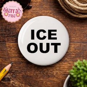 May include: A white, circular badge featuring the black text "ICE OUT". The badge is displayed on a wooden surface, with a pencil, sunglasses, and a small plant in the background.