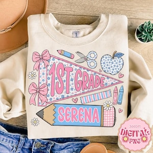 May include: A cream-colored crewneck sweatshirt featuring a graphic print of a pennant flag with the text 1st GRADE, a large pencil with the name SERENA, a floral apple, scissors, and bows. The design includes pink, blue, and white colors.