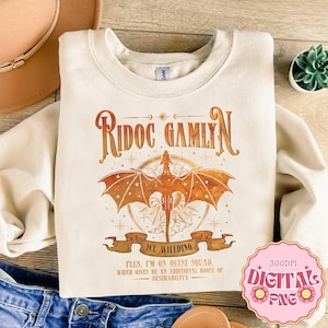 Op de afbeelding: Een crèmekleurig sweatshirt met ronde hals en een afbeelding van een draak met gespreide vleugels boven een banier. De tekst luidt "RIDOC GAMLYN", "ICE WIELDING" en "PLUS, I'M ON QUEST SQUAD, WHICH GIVES ME AN ADDITIONAL BOOST OF DESIRABILITY".
