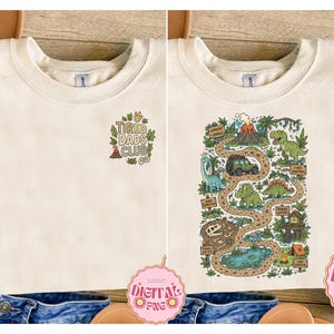 May include: Cream-colored sweatshirt with two designs. One features the text "TIRED DADS CLUB" with a volcano and leaves. The other design is a dinosaur-themed map with a volcano, dinosaurs, and a jeep.