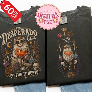 May include: A dark gray t-shirt featuring a graphic of a fluffy cat wearing sunglasses and a crown, holding a cocktail glass. The design includes a treasure chest, skull, daggers, and the text "THE DESPERADO CLUB" and "SO FUN IT HURTS."