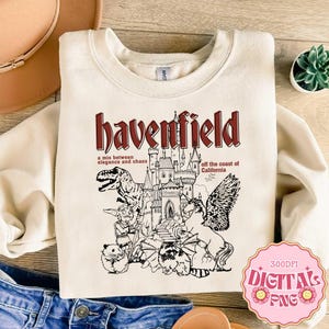May include: A cream-colored crewneck sweatshirt featuring a dark red and black graphic print. The design includes a castle, a dinosaur, a winged horse, a small creature, and a raccoon, with the text "havenfield" and "a mix between elegance and chaos off the coast of California".