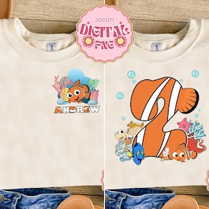May include: Cream-colored t-shirt featuring a cartoon clownfish design with the name "ANDREW" below it. The design includes coral and other sea creatures. The shirt also has a large orange number 2 design with sea creatures.