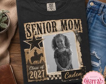 May include: A charcoal gray t-shirt featuring a western-themed graphic with the text SENIOR MOM, Class of 2027, and Caden. The design includes a star, a silhouette of a person on a horse, and a central black and white photograph of a young person.