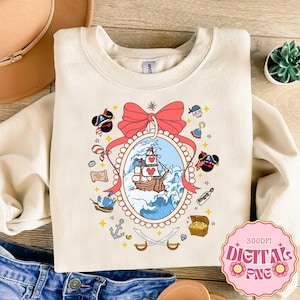 Könnte beinhalten: Cremefarbenes Sweatshirt mit Piratenmotiv. Das Design zeigt ein Schiff, das auf blauen Wellen in einem ovalen Rahmen segelt, umgeben von Piraten-Symbolen wie einer Schatzkiste, Schwertern und einer Schleife. Das Design ist mit roten, blauen und goldenen Details akzentuiert.