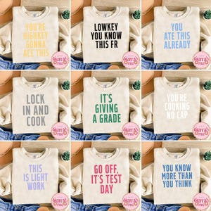 May include: Nine cream-colored sweatshirts with various phrases printed in different colors. The phrases include "YOU'RE HIGHKEY GONNA ACE THIS", "LOWKEY YOU KNOW THIS FR", and "YOU ATE THIS ALREADY". Other phrases are "LOCK IN AND COOK", "IT'S GIVING A GRADE", "YOU'RE COOKING NO CAP", "THIS IS LIGHT WORK", "GO OFF, IT'S TEST DAY", and "YOU KNOW MORE THAN YOU THINK".